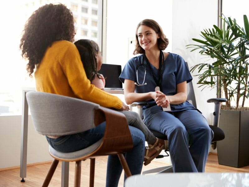 Mother And Daughter Having Consultation With Female Pediatrician Wearing Scrubs In Hospital Office